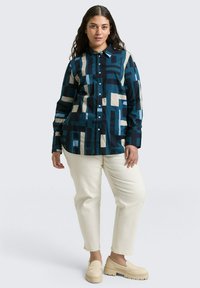 Knoopshirt in teal, marineblauw en crème met geometrisch patroon. Lange mouwen, relaxte pasvorm. Combineren met crème broek en lichtbeige instappers.