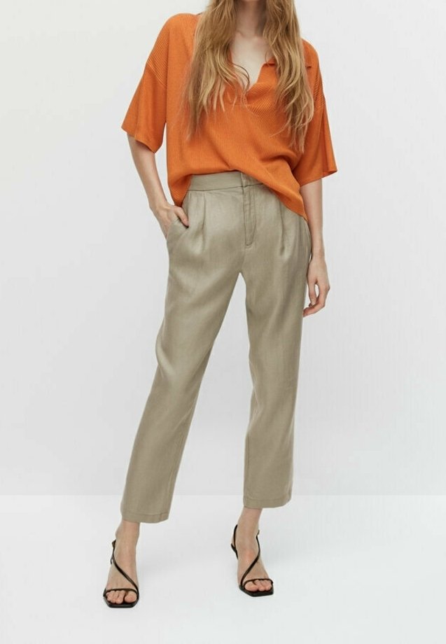 Femme portant un chemisier orange à manches courtes, un pantalon beige taille haute et fuselé, ainsi que des sandales à talons noires avec brides, debout avec une main dans la poche.