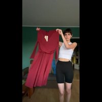 Dolga burgujska obleka z globokim V-izrezom, dolgimi rokavi in zategnjenim pasom; gladka tekstura; drži jo oseba v belem kratkem topu in črnih hlačah.