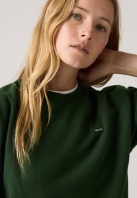 Sweatshirt vert foncé avec un col rond, arborant un petit logo noir Levi's sur la poitrine. Texture douce et coupe décontractée.