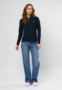 Marineblå langermet polo skjorte med tre knapper i placketen og brodert logo, sammen med lyseblå bell-bottom jeans og hvite joggesko.