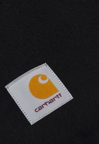 Svart stoff med teksturert utseende, med et kvadratisk hvitt merke som viser en gul logo og ordet "Carhartt" i rødt.