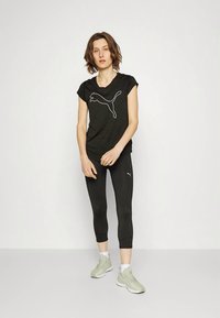 Černé sportovní tričko s krátkými rukávy zdobí bílý logo Puma, doplněné černými legínami a světle šedými běžeckými botami. Celkově hladké textury.