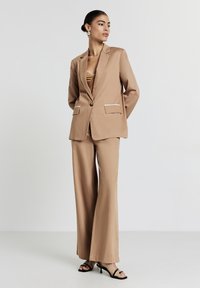 Blazer beige ajusté avec des détails de poche blancs, associé à un pantalon large. Tissu lisse, coupe structurée et design minimaliste.