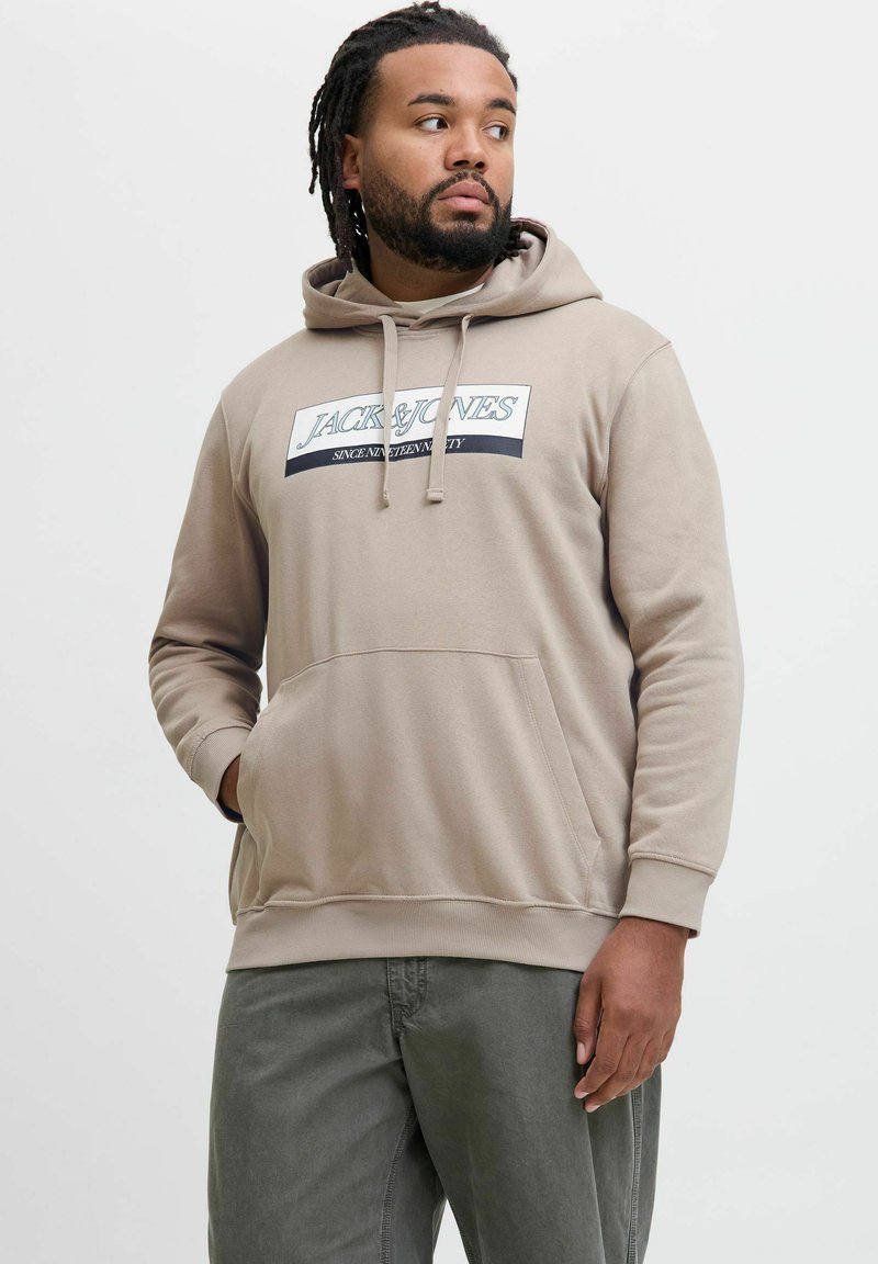 Beige hoodie lavet af blødt stof med en front kangaroo-lomme, snøre-hætte og et trykt logo på brystet. Kombineret med grønne bukser.
