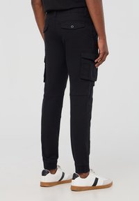 Czarne spodnie cargo z elastycznymi mankietami, kieszeniami bocznymi i tylnymi kieszeniami z klapką, wykonane z solidnego materiału. W połączeniu z białymi trampkami.