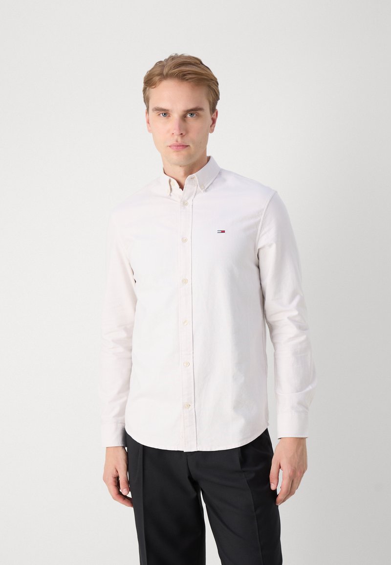 Camisa blanca de botones hecha de algodón, con mangas largas, un cuello puntiagudo y un pequeño logo cerca del pecho. Combinada con pantalones negros.
