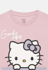 T-shirt rosa de mangas curtas com um grande gráfico da Hello Kitty, um laço lavanda e o texto "Good vibes" em branco cursivo. Material de algodão macio.