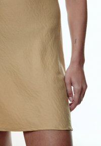 Lichtbeige mouwloze jurk met een gestructureerde afwerking, met een zoom die boven de knie valt. Hand rust nabij de rand van de rok.
