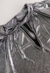 Gros plan de tissu métallique argenté brillant avec une découpe en forme de trou de serrure et des coutures plissées près du col sur un vêtement.