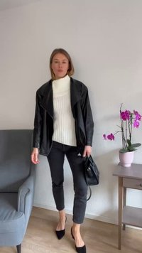 Schwarze Kunstlederjacke über einem weißen, gerippten Pullover, kombiniert mit schwarzen Slim-Fit-Hosen und spitzen schwarzen Absätzen. Eine kleine schwarze Tasche wird getragen.