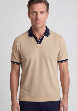 Man met kort bruin haar, gekleed in een beige poloshirt met een donkerblauwe kraag en mouwranden, staand tegen een effen lichte achtergrond.