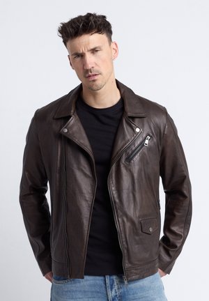 Mann mit kurzen Haaren, der eine dunkelbraune Lederjacke über einem schwarzen Shirt und blauen Jeans trägt, steht mit den Händen in den Taschen vor weißem Hintergrund.