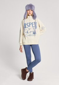 Beżowa bluza z grafiką "ASPEN Colorado", niebieskie legginsy i brązowe botki za kostkę. Zawiera fioletowy futrzany kapelusz z klapkami na uszy.