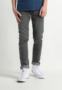 Jeans grigi slim-fit con una texture morbida, orli risvoltati, abbinati a sneakers alte bianche con dettagli rossi.