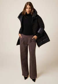 Giacca puffer nera con cappuccio, indossata su una maglia nera, abbinata a pantaloni larghi in tessuto marrone e tacchi neri a punta.