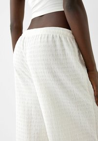 Bílé široké kalhoty s texturou a elastickým pasem. Látka má zmačkaný vzor a jemný vzhled.