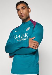 Pitkähihaine turkoosi urheilu paita, jossa on violetti olkapääkorostus. Ominaisuuksiin kuuluu korkea kaulus ja valkoinen logo, jossa on etupuolella "QATAR AIRWAYS" teksti.