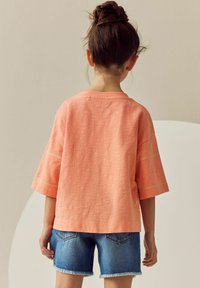 Korallfärgad oversized T-shirt med korta ärmar och dekorativa sömmar på ärmarna, kombinerad med denimshorts med fransade fållar.