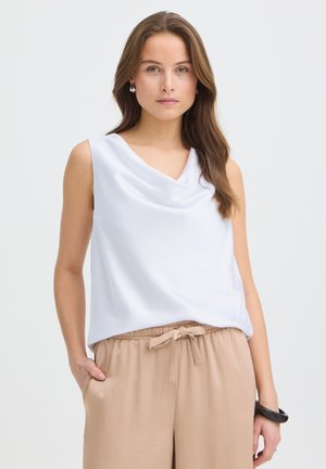 Jeune femme aux longs cheveux bruns portant un haut blanc sans manches drapé et un pantalon beige à cordon, posant devant un fond uni.