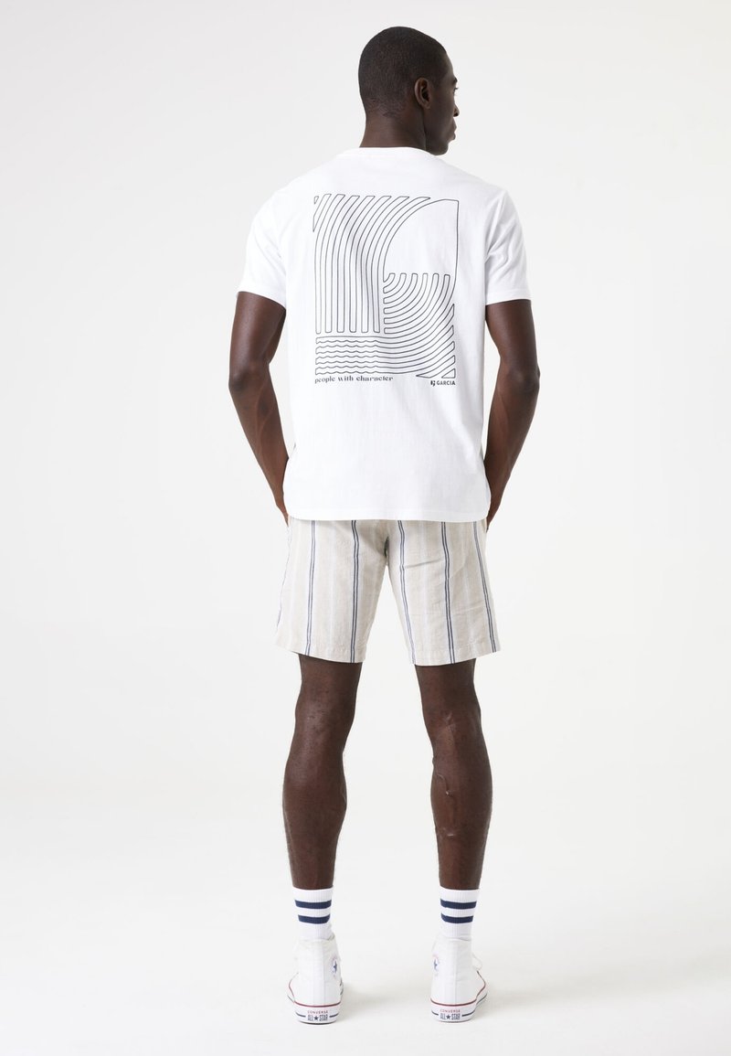 Camiseta blanca de algodón con un gráfico de línea negra en la parte posterior, combinada con pantalones cortos a rayas beige, calcetines blancos y zapatillas blancas.