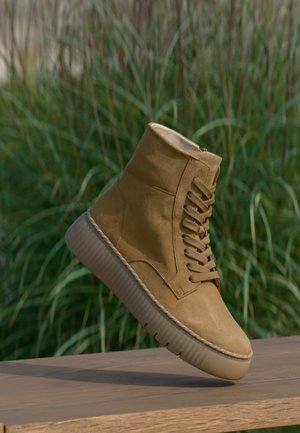 Botte montante en daim marron clair avec une épaisse semelle en caoutchouc reposant sur une surface en bois avec des plantes vertes en arrière-plan.