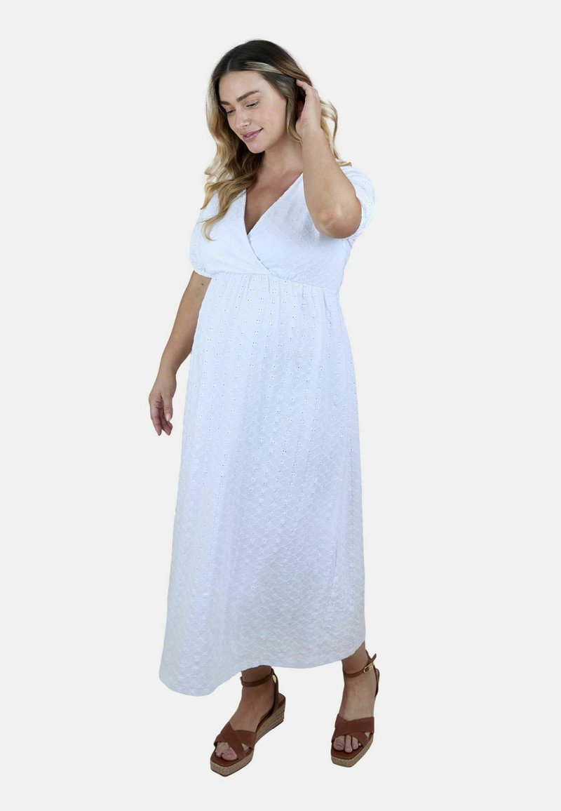 Egy fehér texturált wrap ruhát viselő kismama, barna ékcipővel, haját simogatva, egyszínű háttér előtt áll.