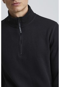 Crna zip-up dukserica s visokim ovratnikom, s kratkim zatvaračem i glatkom, mekanom teksturom. Minimalistički dizajn bez vidljivih uzoraka.