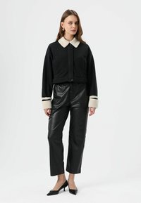 Zwarte cropped jas met een crèmekleurige nepbontkraag, gecombineerd met hooggetailleerde zwarte nepleren broek en zwarte pumps met spitse neus.