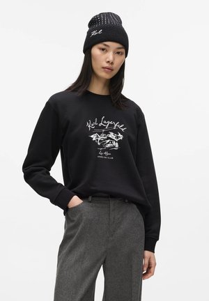 Kvinde iført en sort hue og sort sweatshirt med print af bjerg og ski-lift, kombineret med grå uldbukser, poserer med den ene hånd i lommen.