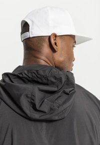 Giacca nera con cappuccio e tessuto arricciato al collo, indossata con un berretto bianco visibile dal retro, che mette in evidenza un design urbano slanciato.