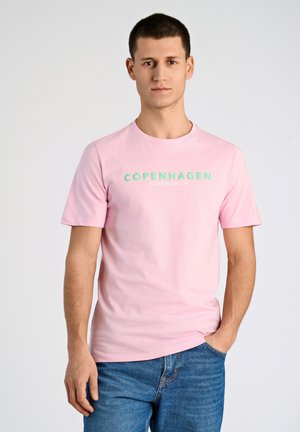 Camiseta de algodón rosa con las palabras "COPENHAGUE" y "LINDBERGH" estampadas en verde menta. Mangas cortas y cuello redondo. Combinada con jeans azules.