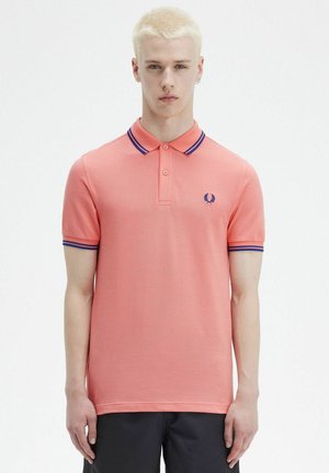 Polo coral con cuello a rayas azules y moradas, de mangas cortas y un logo bordado en el lado izquierdo del pecho. Textura de algodón suave.