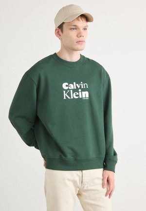Sweatshirt vert avec le logo "Calvin Klein" blanc, col rond et manches côtelées. Le modèle porte une casquette de baseball beige. Arrière-plan clair.