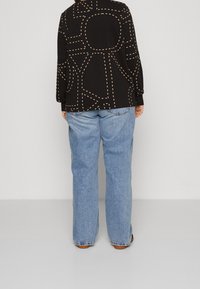 Svart långärmad topp med ett geometriskt beige prickmönster; ljusblå raka jeans; avslappnat fotklädsel synligt längst ner.