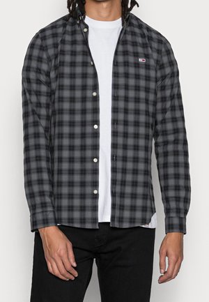 Camisa a cuadros en negro y gris con cuello abotonado, botones blancos y logotipo. Uso en capas sobre una camiseta blanca, confeccionada en tela tejida.