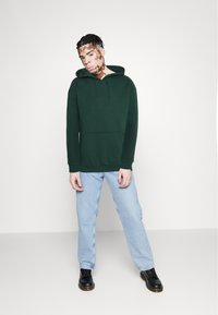 Groene katoenen hoodie met een voorkantzak, trekkoordcapuchon en geribbelde manchetten, gecombineerd met lichtblauwe denim jeans en zwarte veterschoenen.