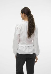 Camicia bianca con maniche lunghe a sbuffo, orlo arricciato in vita e collo alto. Abbinata a pantaloni neri; tessuto liscio con texture minima.
