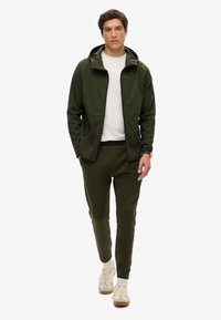 Felpa verde con zip e cappuccio, abbinata a pantaloni jogger coordinati. Maglietta bianca sotto, completata con sneakers chiare e calzini al polpaccio.