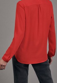 Blusa rossa a maniche lunghe in tessuto liscio con una leggera lucentezza; presenta un orlo arrotondato e polsini con bottoni. Indossata con jeans scuri.