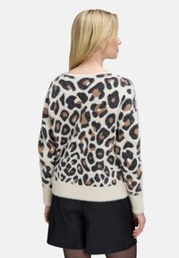 Žena s rovnými blond vlasmi, oblečená v huňatej svetri s leopardím vzorom a čiernych šortkách, otočená chrbtom k jednému pozadiu.