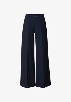 Tumši zili plataji bikses ar priekšpuses krokām, jostas cilpām un pogas aizdari uz baltas virsmas.