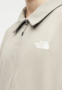 Μπεζ μπουφάν με γιακά και φερμουάρ μπροστά, με άσπρο κεντημένο λογότυπο "The North Face" στο στήθος.