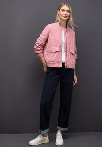 Giovane donna che indossa una giacca bomber rosa, una T-shirt bianca, jeans scuri arrotolati e sneaker bianche, in piedi contro un muro grigio.