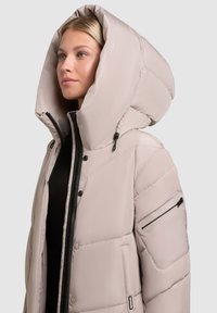 Kvinde iført en lys beige pufferjakke med en overdimensioneret hætte og en sort lynlås, der ser lidt til siden på en ensfarvet baggrund.