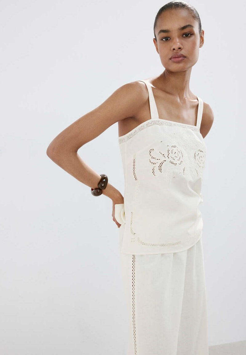 Jeune femme portant un haut blanc sans manches avec une broderie florale en découpe et un pantalon large assorti, posant avec la main sur la hanche.