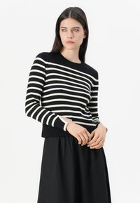 Femme aux cheveux bruns portant un pull à rayures horizontales noir et blanc et une jupe noire, se tenant devant un fond blanc.
