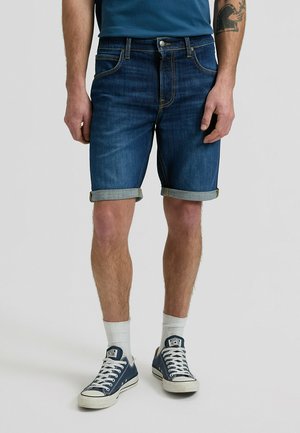 Hombre con pantalones cortos de mezclilla oscuros arremangados, calcetines blancos hasta el tobillo, zapatillas bajas azul marino y una camiseta azul, de pie con los brazos relajados.