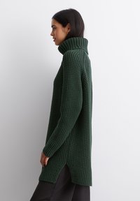 Grøn strikket sweater med høj krave, der har et struktureret mønster og sidespalter. Ærmerne er lange og tætsiddende.