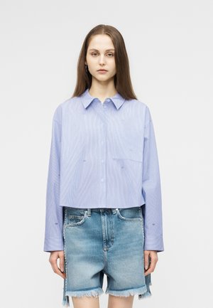 Sieviete ar gariem brūniem matiem valkā gaiši zilu pārāk lielu pogām aizpogājamu kreklu un izbalējušas augsta vidukļa džinsa šortus ar saplēstām malām.